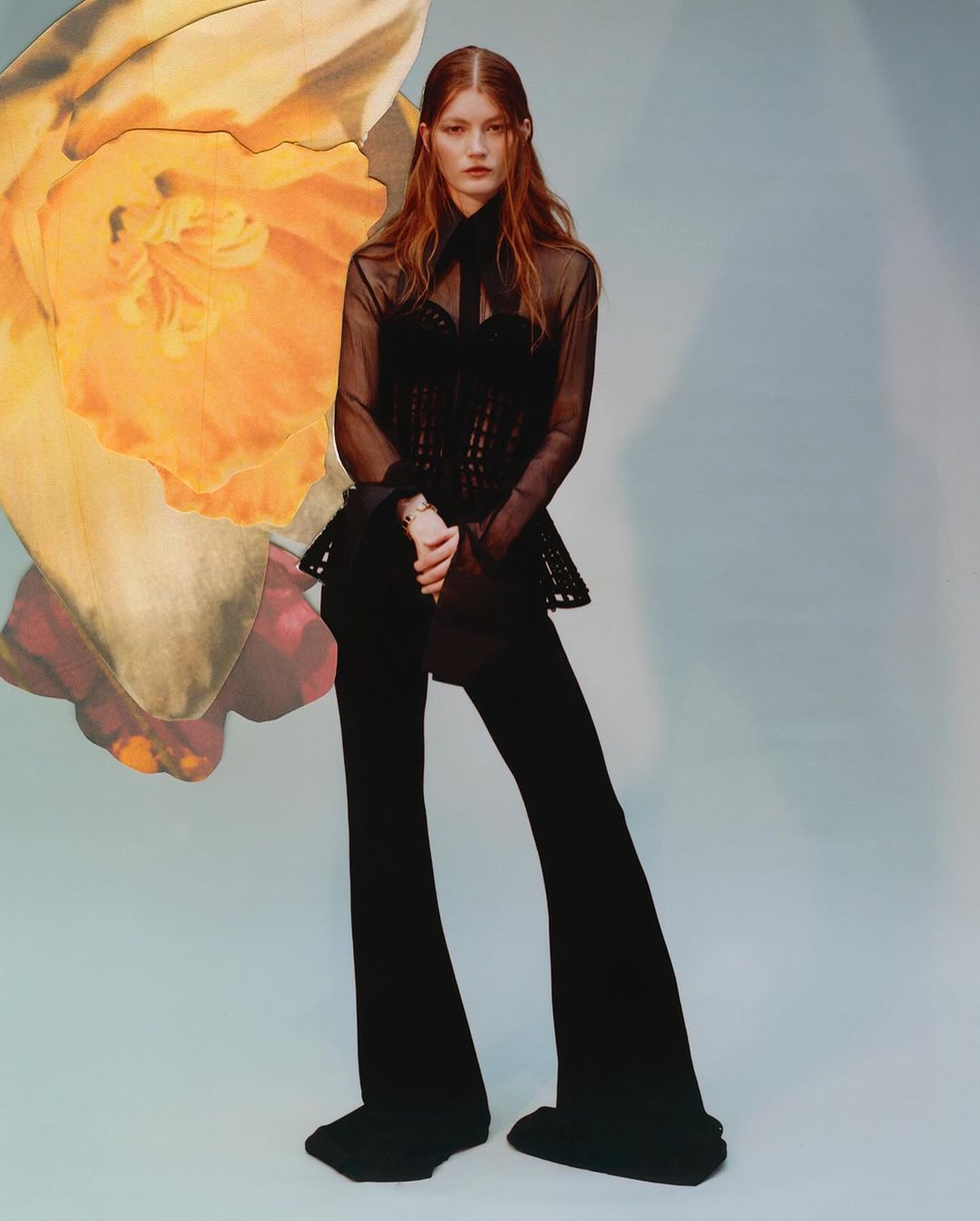 A red-haired fashion model poses in a sheer black blouse and black trousers against a light blue background with a yellow flower.