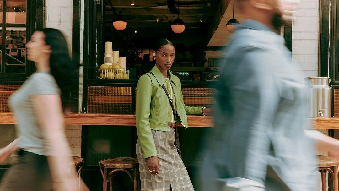 Woman in a green jacket and plaid skirt stands in front of an outdoor cafe, with blurred figures walking by on a city sidewalk.