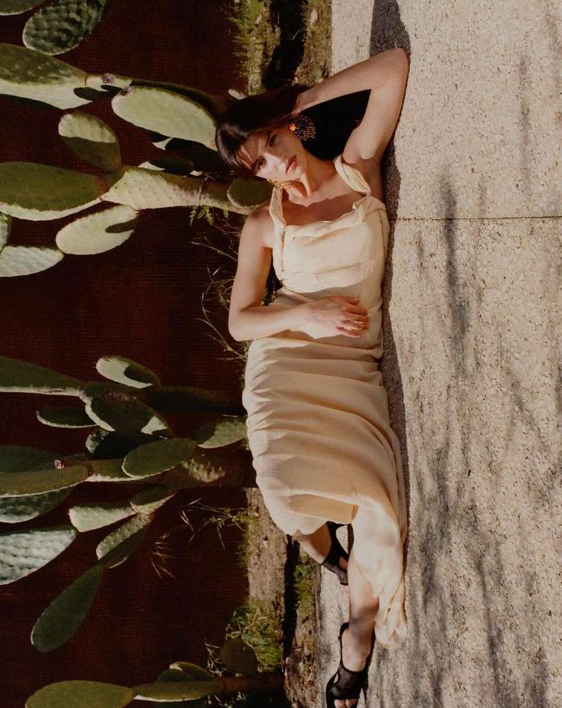 Woman in cream sleeveless dress reclining outdoors against large prickly pear cacti in direct sunlight