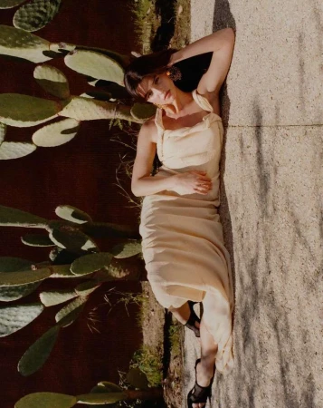 Woman in cream sleeveless dress reclining outdoors against large prickly pear cacti in direct sunlight