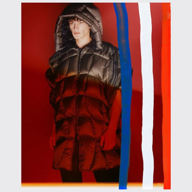 Adult male poses in a hooded, sleeveless, black and red puffer jacket against a red backdrop with vertical blue, white, and orange paint strokes.