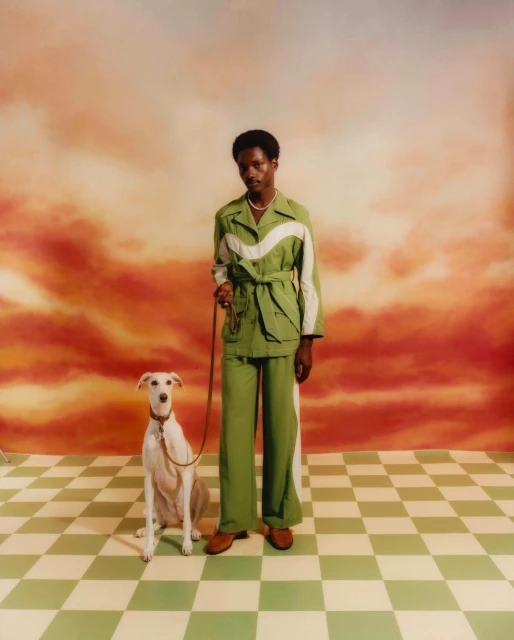 Elegant man in green suit poses with white greyhound dog in a studio against a scenic orange and white cloud background.