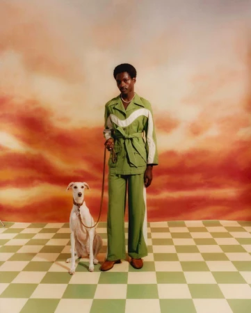 Elegant man in green suit poses with white greyhound dog in a studio against a scenic orange and white cloud background.