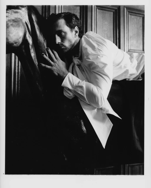 Monochrome portrait of a man in a white ruffled shirt reclining on a dark horse sculpture against paneled walls, striking a dramatic pose.
