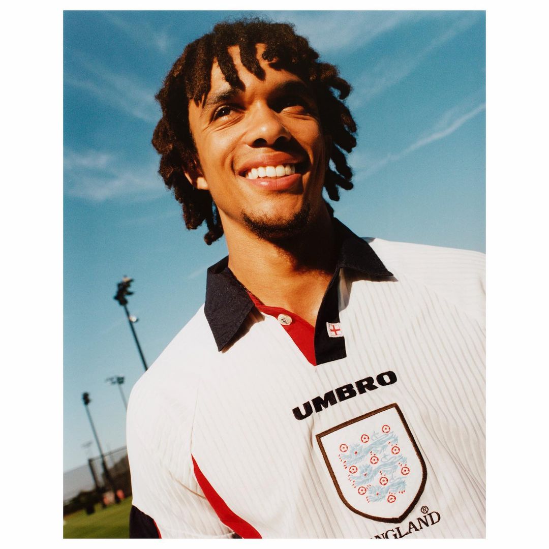 Portrait of a smiling adult male wearing an England soccer jersey against a blue sky backdrop with outdoor sport field elements.