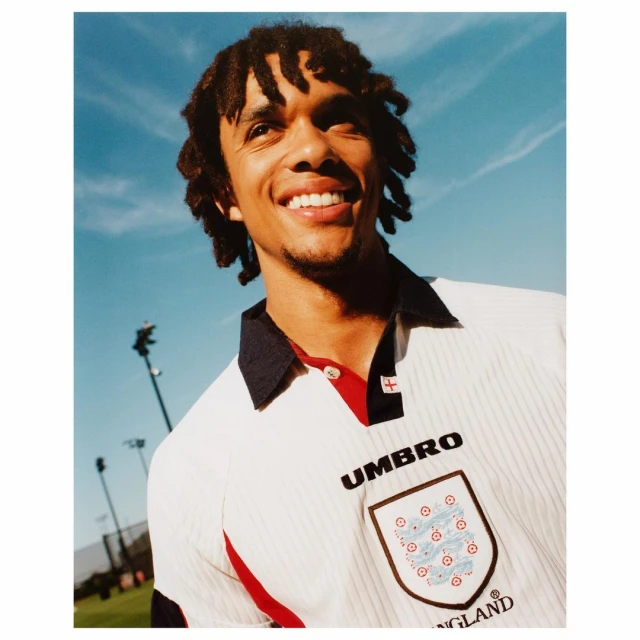 Portrait of a smiling adult male wearing an England soccer jersey against a blue sky backdrop with outdoor sport field elements.