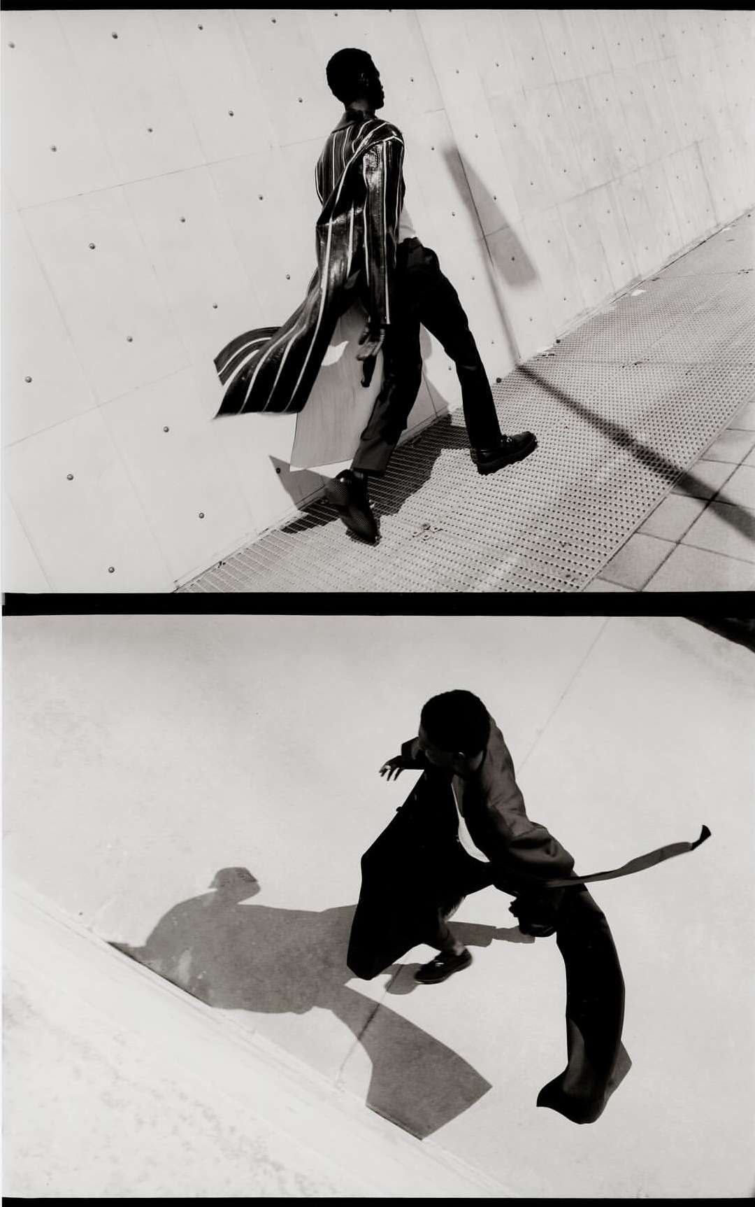 Monochrome fashion shot of a Black male model in a long striped coat and black pants, walking in an urban setting against a concrete wall.