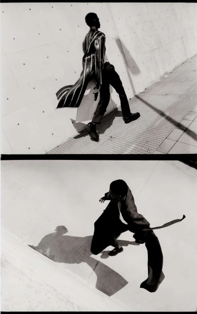 Monochrome fashion shot of a Black male model in a long striped coat and black pants, walking in an urban setting against a concrete wall.