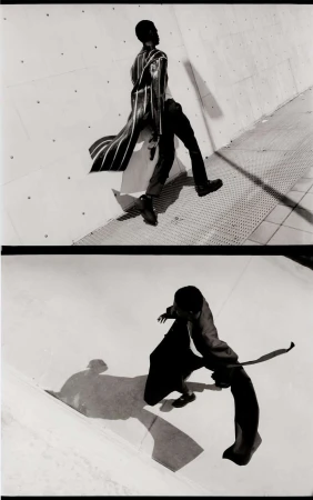 Monochrome fashion shot of a Black male model in a long striped coat and black pants, walking in an urban setting against a concrete wall.