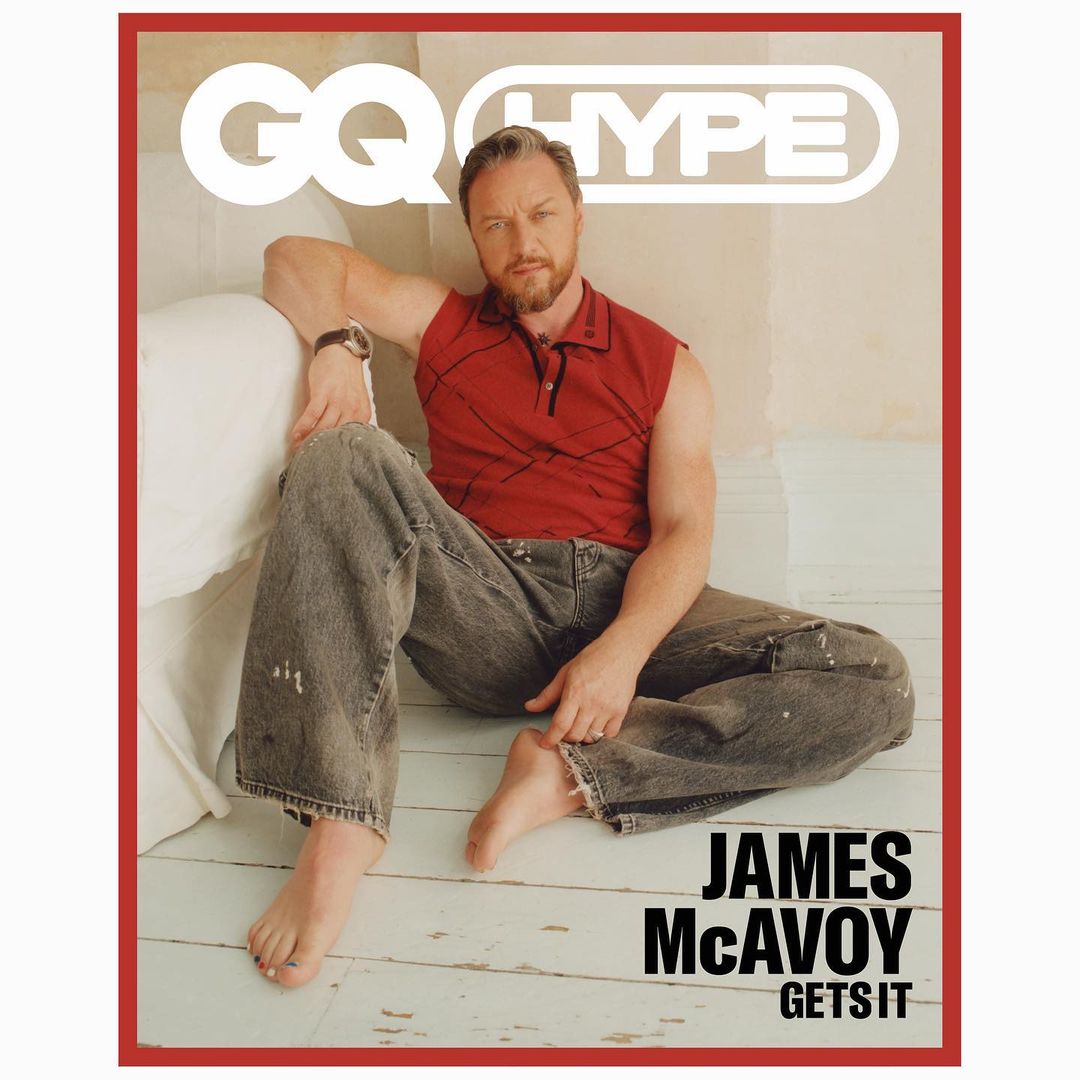 James McAvoy on GQ Hype cover: The adult male celebrity sits cross-legged on the floor wearing a red shirt and grey jeans in a studio setting.