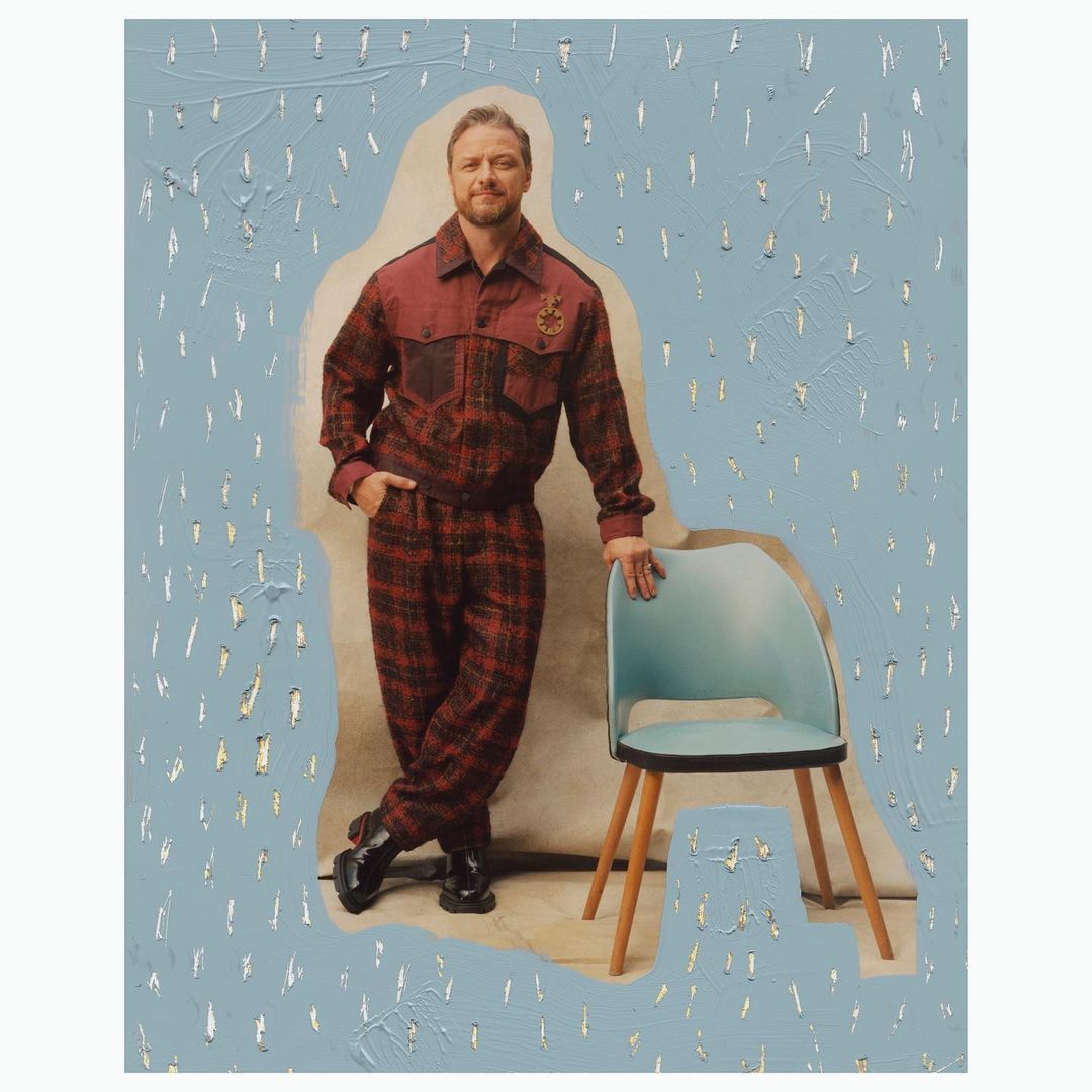 Portrait of a man wearing a red plaid suit leaning against a light blue chair in front of a painted backdrop with light blue and silver.
