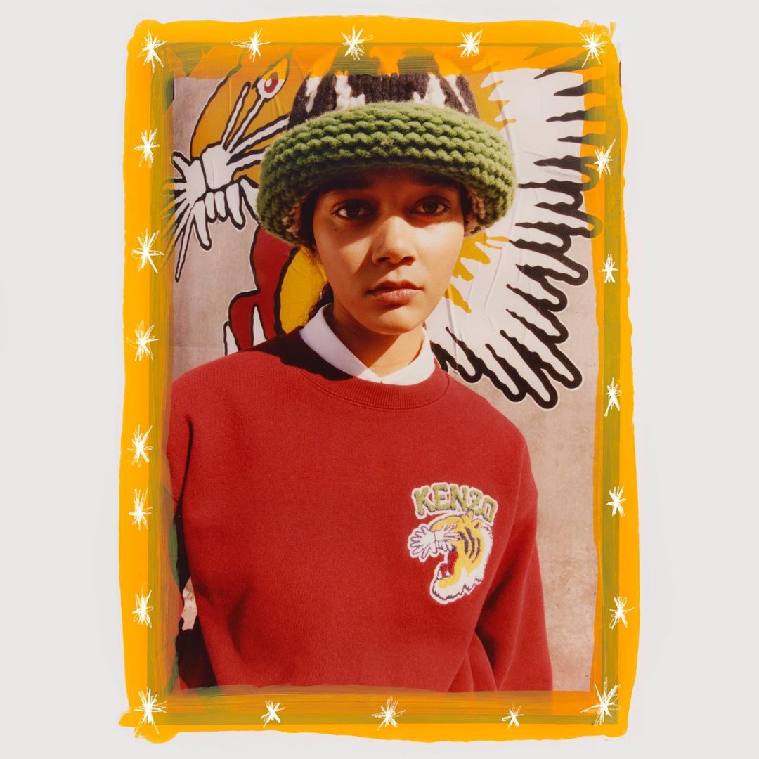 Fashion shot of person in rust-colored sweatshirt and green knitted bucket hat, posing in front of a graphic background with a yellow border.