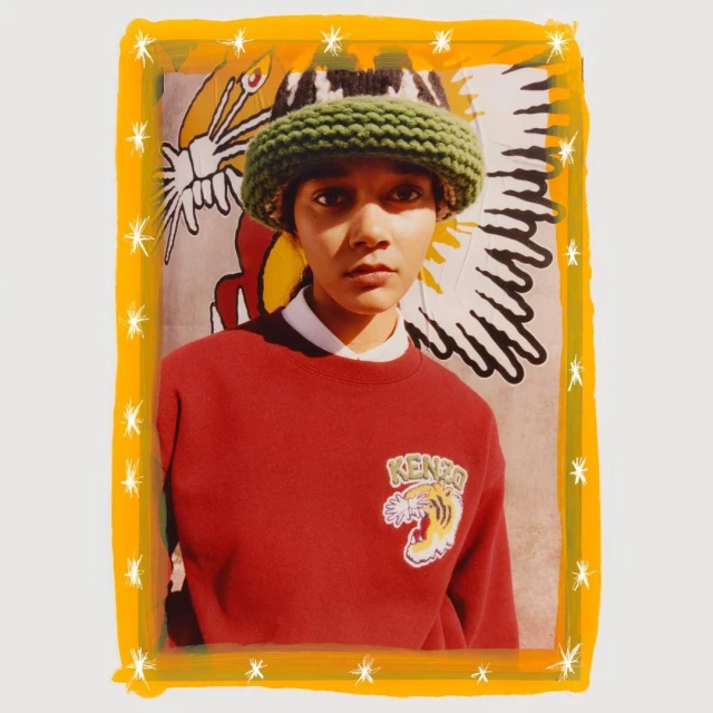 Fashion shot of person in rust-colored sweatshirt and green knitted bucket hat, posing in front of a graphic background with a yellow border.