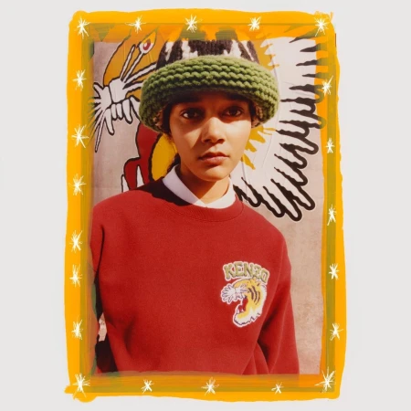 Fashion shot of person in rust-colored sweatshirt and green knitted bucket hat, posing in front of a graphic background with a yellow border.