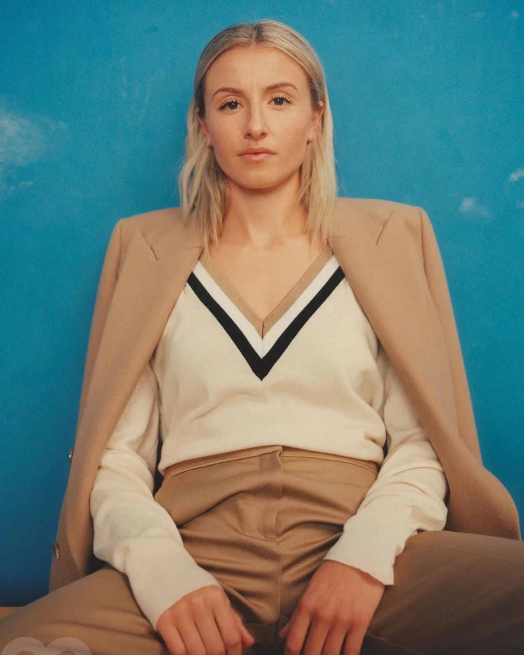 Blonde woman in a beige blazer and pants poses against a blue studio backdrop with faint clouds, creating a modern and fashionable portrait.