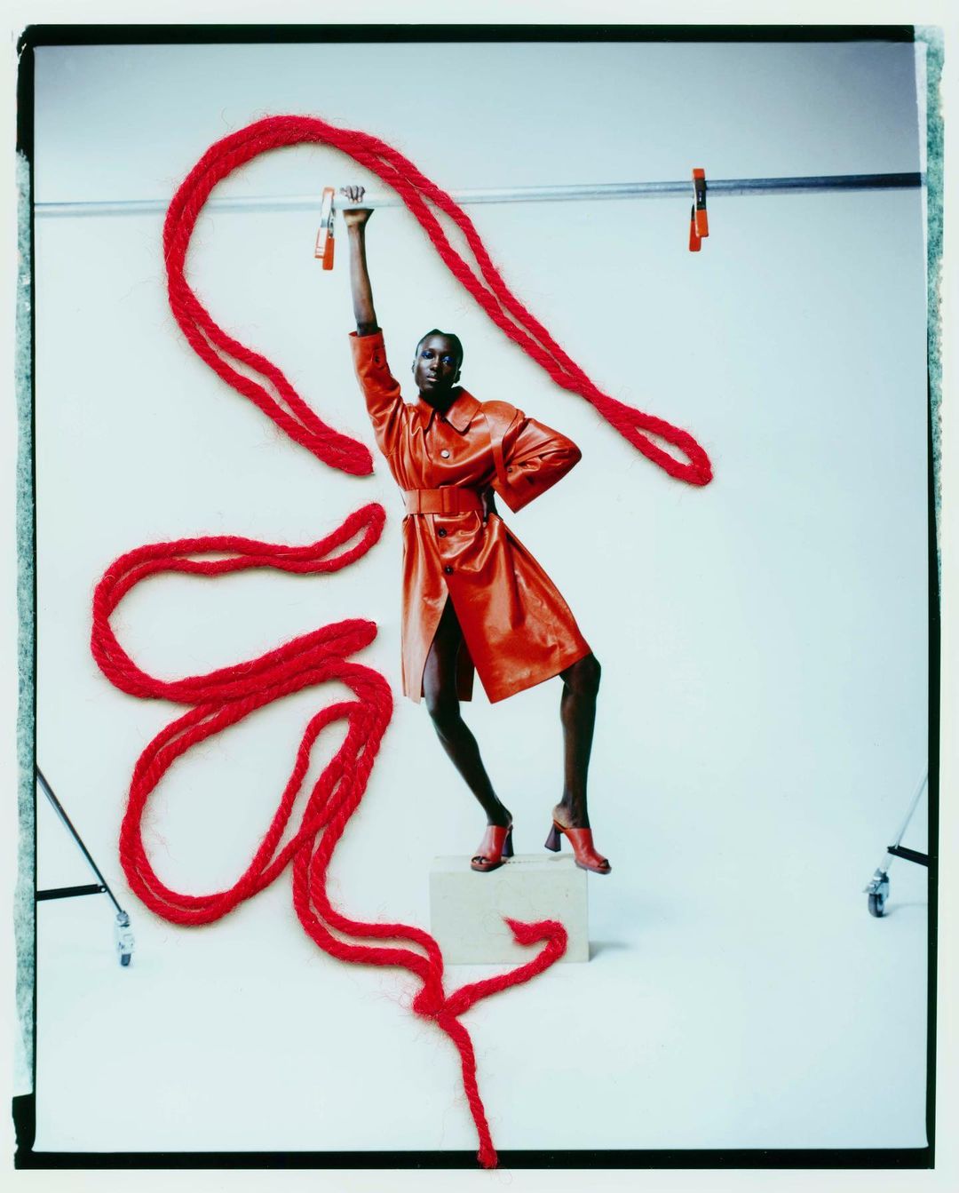 Editorial fashion shot of a Black woman in an orange coat and heels, standing on a cube, holding red yarn in a photo studio.