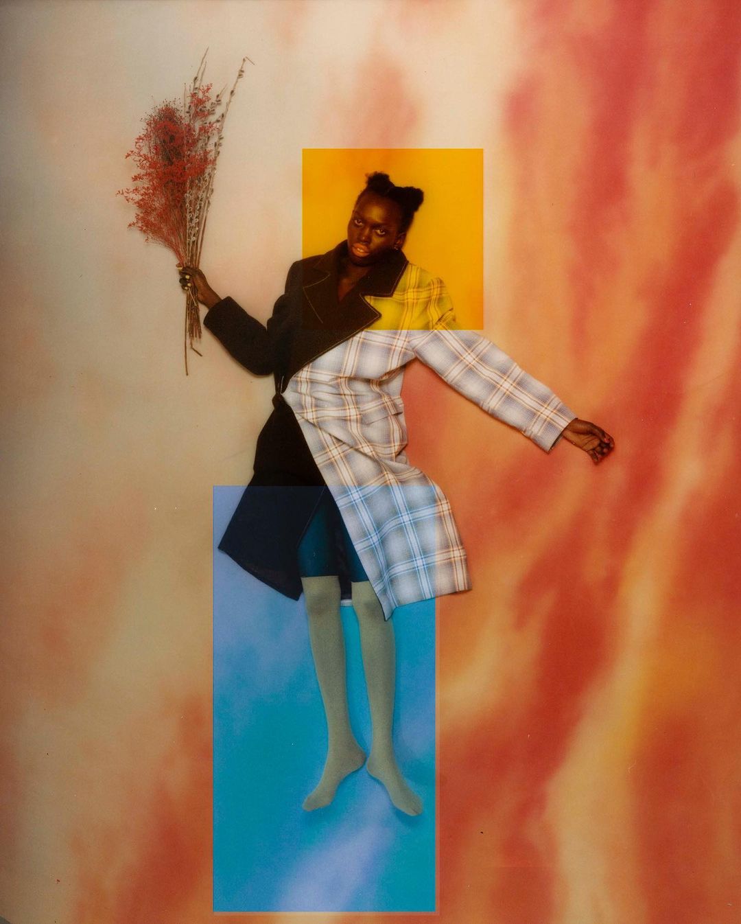 Elegant African American woman in a patchwork blazer and neutral-tone tights holds red foliage against an orange and blue background.
