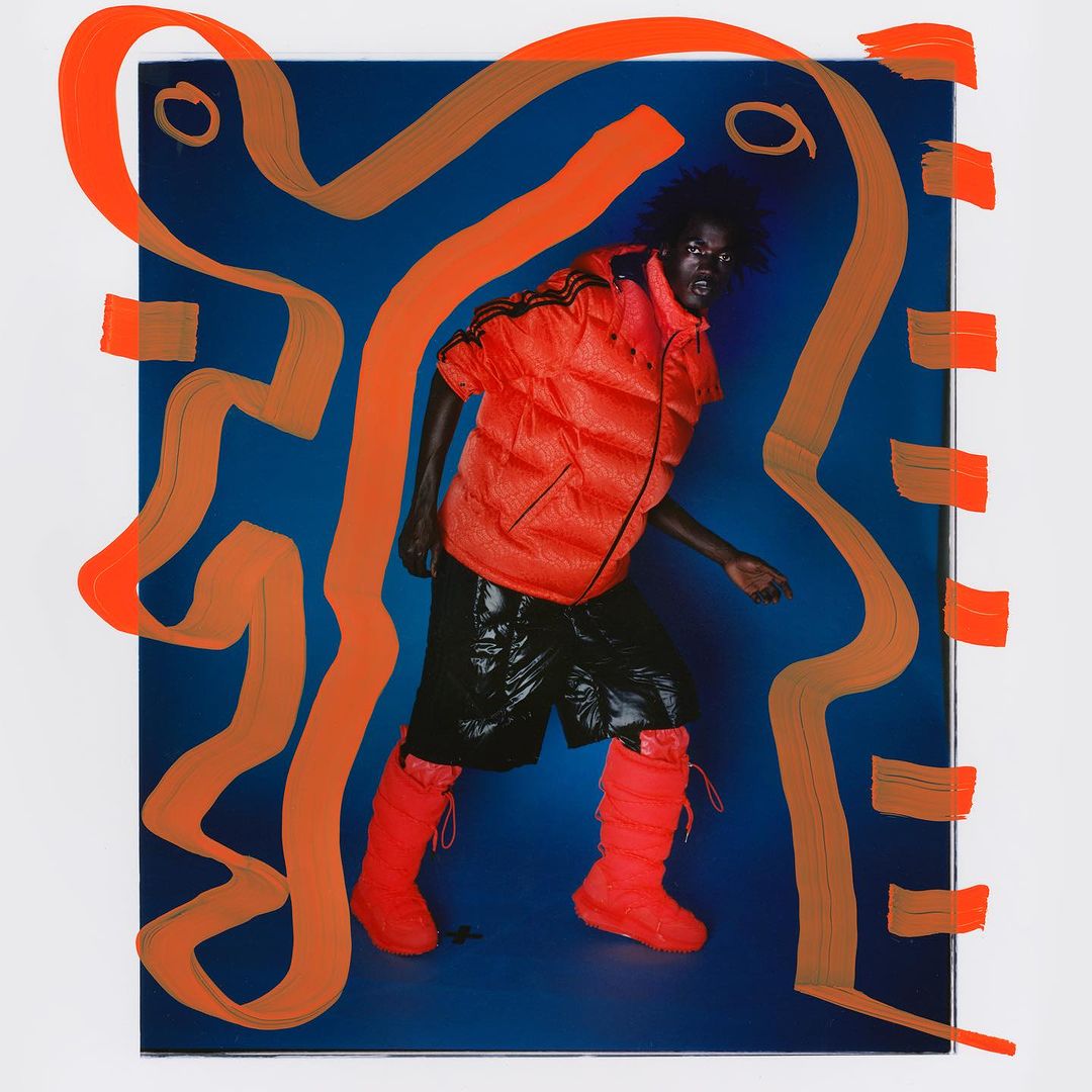 Fashion shot of person in orange puffer vest, black shorts, and orange boots against blue backdrop with orange abstract shapes.