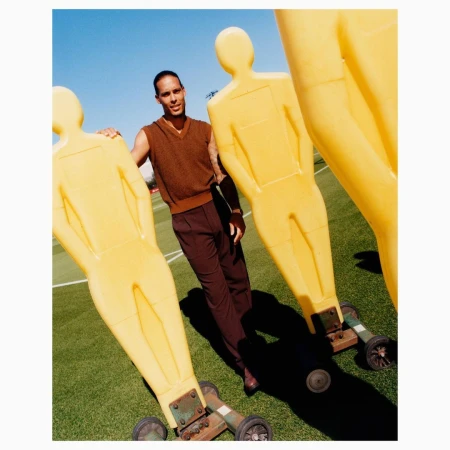 A man in brown sleeveless sweater and trousers poses beside yellow plastic training dummies on green turf under a clear blue sky.