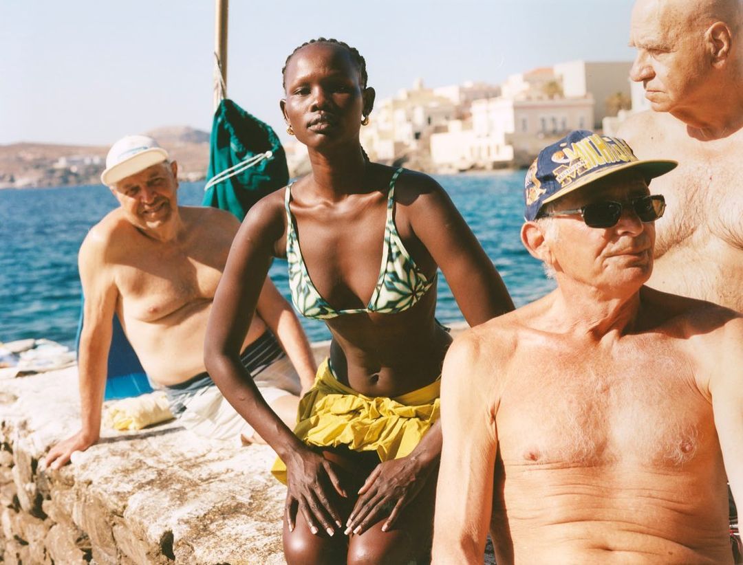 Outdoor gathering featuring a young Black woman in a bikini with older men near the ocean and buildings in the background on a sunny day.