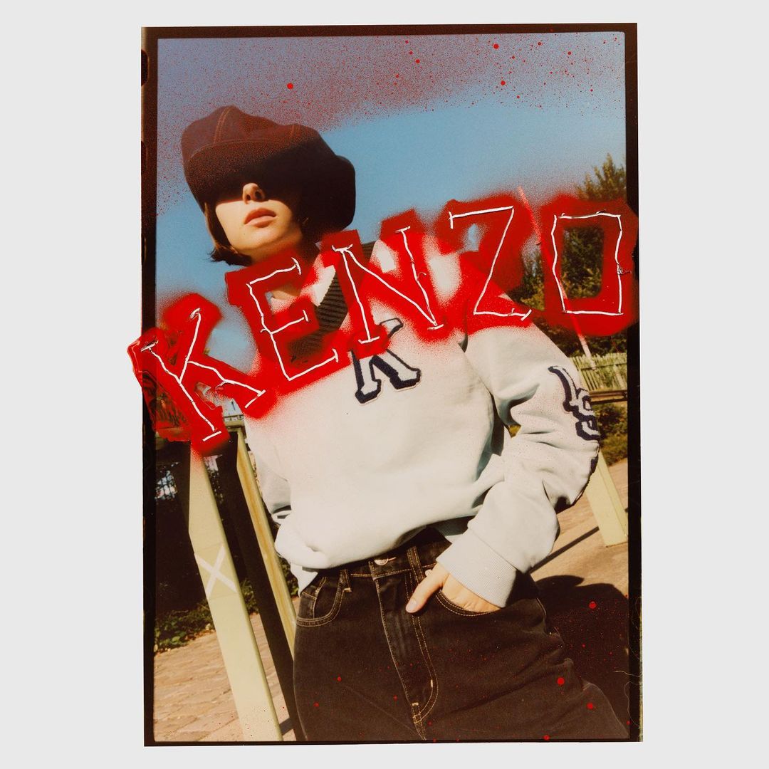 Fashion shot of a woman in a beret, sweater, and jeans, posing outdoors with the word KENZO spray-painted in red across the image.