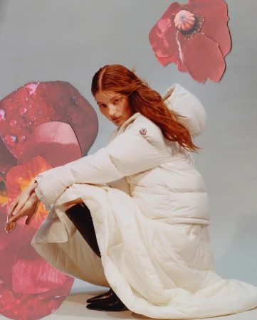 Red-haired female model crouching in a white puffer jacket and matching skirt against a grey background with red floral cutouts