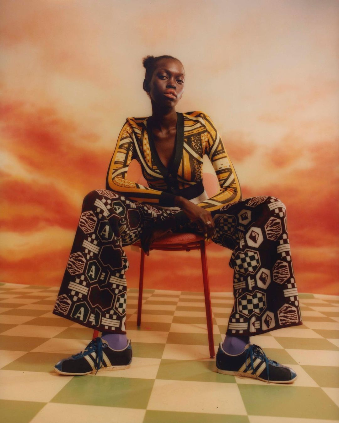 Fashion shot of a Black female model wearing patterned yellow and black clothing, seated on a red chair against a cloudy sky background.