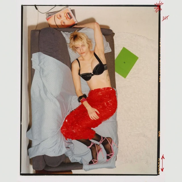 Overhead shot of a blonde woman in a black bra and red sequined skirt reclining on a bed with magazine, laptop, and black stockings and heels.