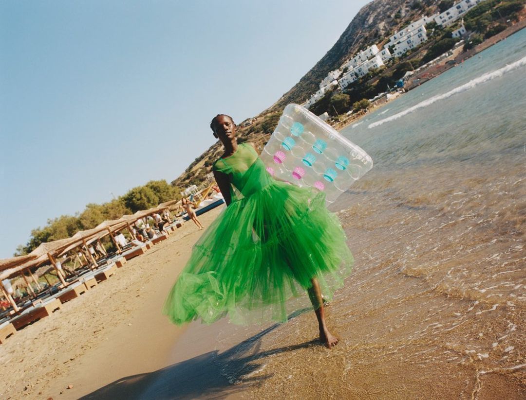 Woman in green tulle dress walks on beach, carrying inflatable pool float, ocean backdrop, sunny day, angled perspective.