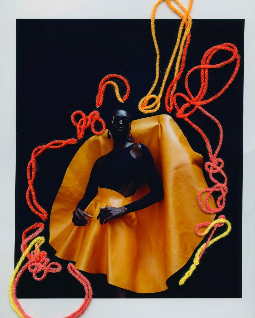 Striking fashion portrait of a black woman in a yellow dress, posing against a black background, with orange yarn art accents.