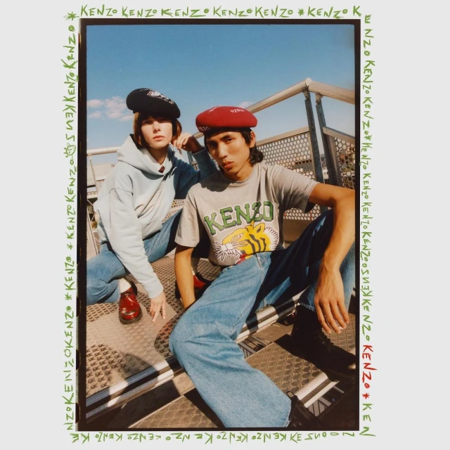 Two fashion models wearing Kenzo apparel and berets pose on metal steps against a blue sky, framed by a Kenzo logo border.