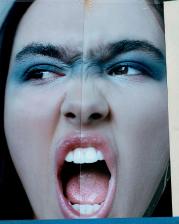 Extreme close-up of a person's face with heavy blue eye makeup, bared teeth, and an open mouth, expressing a strong emotion.