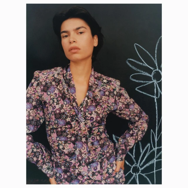 Portrait of a woman in a floral top posing in front of a black wall with white chalk drawings of flowers, studio shot.