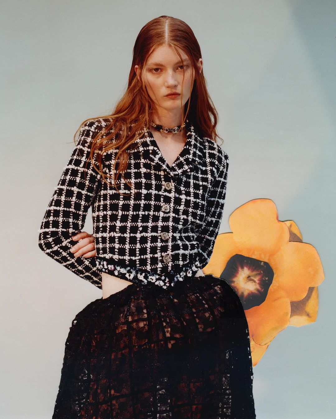 A red-haired woman poses in a black and white jacket and black lace skirt against a light blue background with a paper flower.