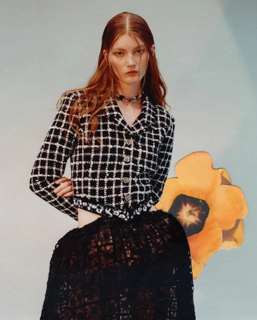 A red-haired woman poses in a black and white jacket and black lace skirt against a light blue background with a paper flower.