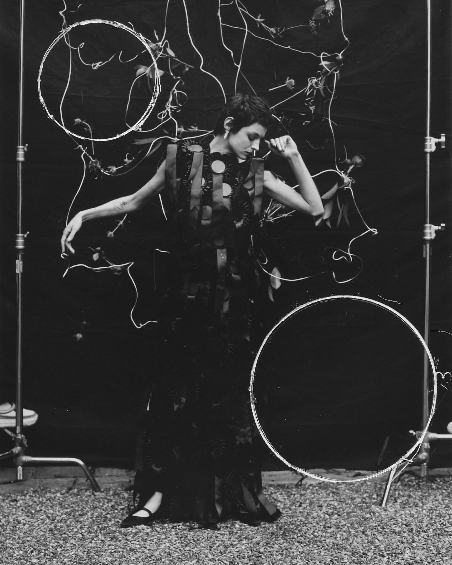 A fashionable adult female model in a dark dress poses in a studio setting with decorative hoops, shot in black and white with soft lighting.