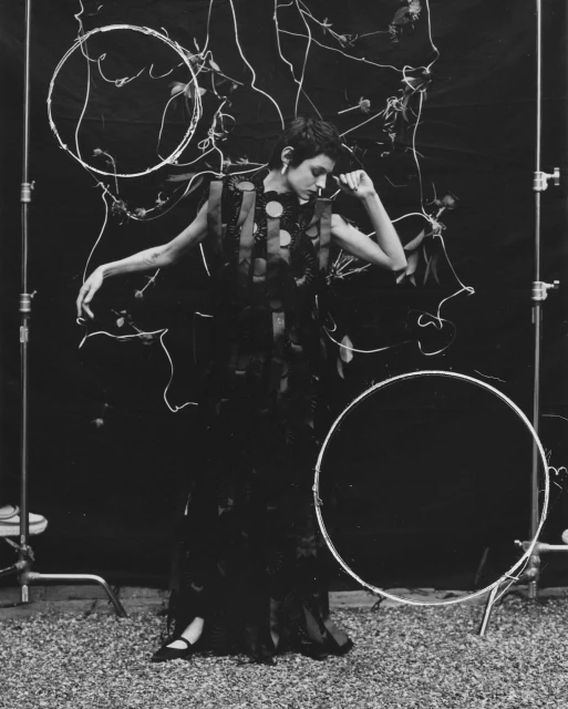 A fashionable adult female model in a dark dress poses in a studio setting with decorative hoops, shot in black and white with soft lighting.