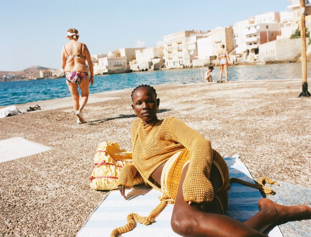 A Black woman lounges on a striped towel at the beach in a yellow knit outfit, with other beachgoers and Mediterranean architecture in the background.