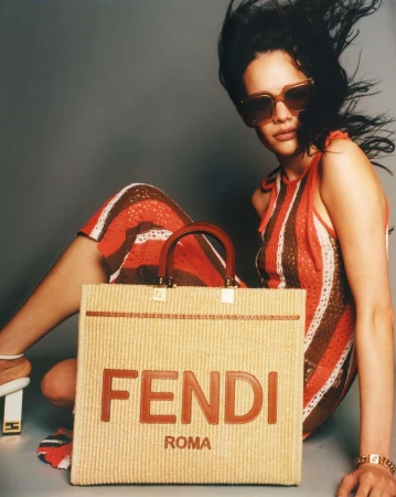 Model wearing sunglasses and striped dress posing behind a large woven Fendi Roma tote bag in a studio setting