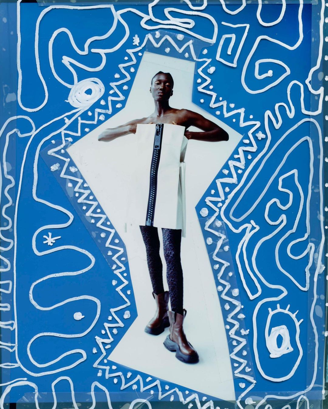 An African-American woman poses in a studio wearing a white dress with a black zipper, black leggings, and brown boots, against a blue background.