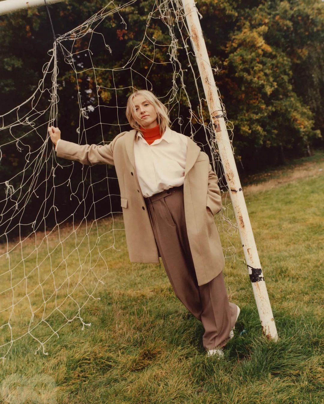 Adult female in tan coat and brown pants poses by soccer goal, outdoors in grassy area with trees; editorial fashion portrait.