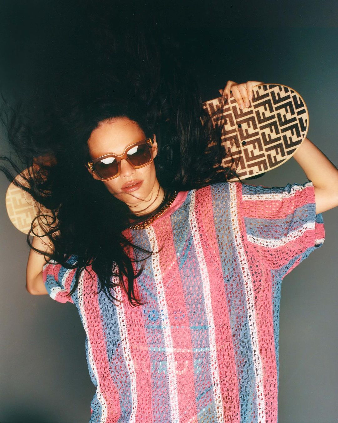 A woman with dark hair and sunglasses poses against a grey background, wearing a striped top and holding a patterned handbag overhead.