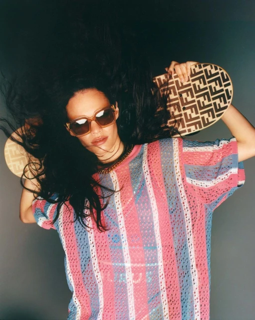 A woman with dark hair and sunglasses poses against a grey background, wearing a striped top and holding a patterned handbag overhead.