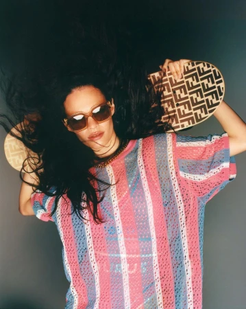 A woman with dark hair and sunglasses poses against a grey background, wearing a striped top and holding a patterned handbag overhead.