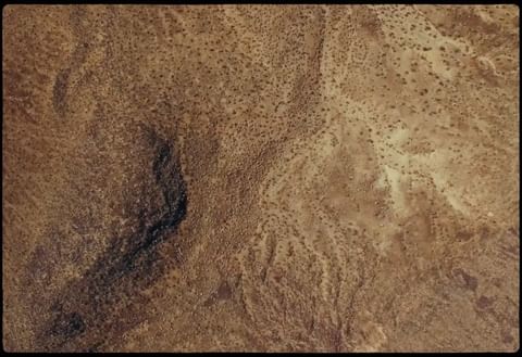 Aerial view of arid desert landscape with rocky terrain, sparse vegetation, and meandering dry riverbed in varying shades of brown.