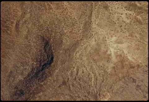Aerial view of arid desert landscape with rocky terrain, sparse vegetation, and meandering dry riverbed in varying shades of brown.