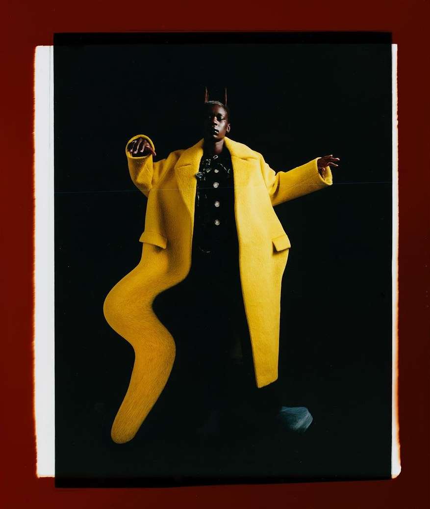 A woman in a vibrant yellow coat poses against a dark backdrop, showcasing a blend of fashion and abstract visual elements.