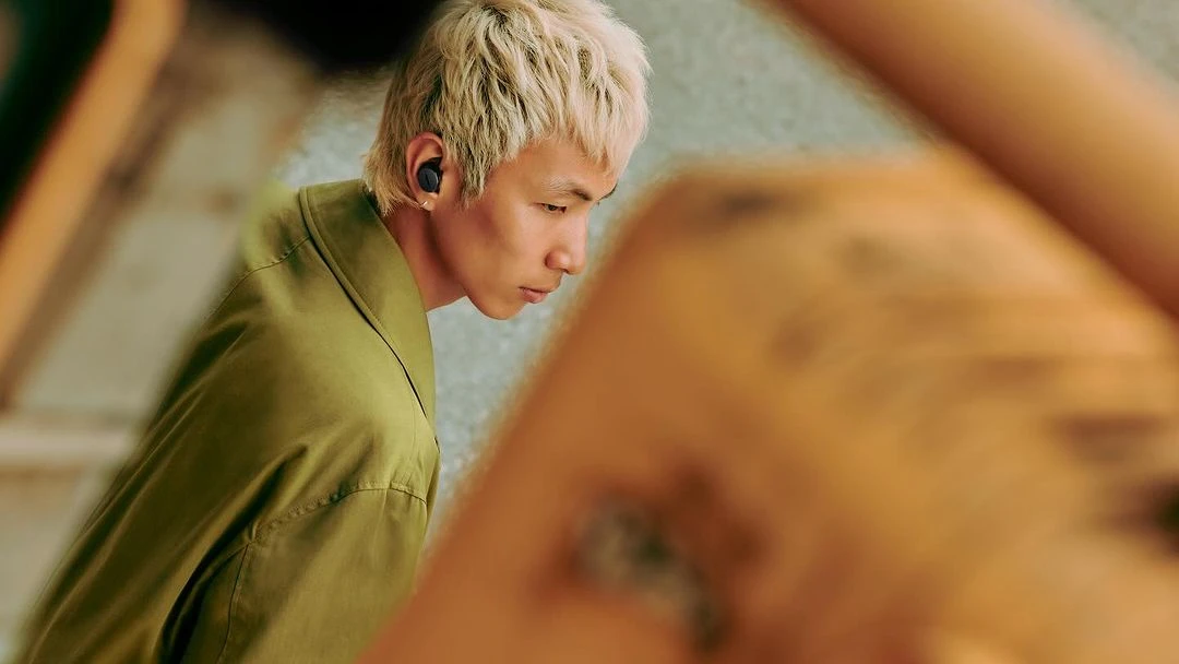 Asian man with blond hair wearing wireless earbuds and an olive-green shirt, gazing down in a close-up shot with blurred background.