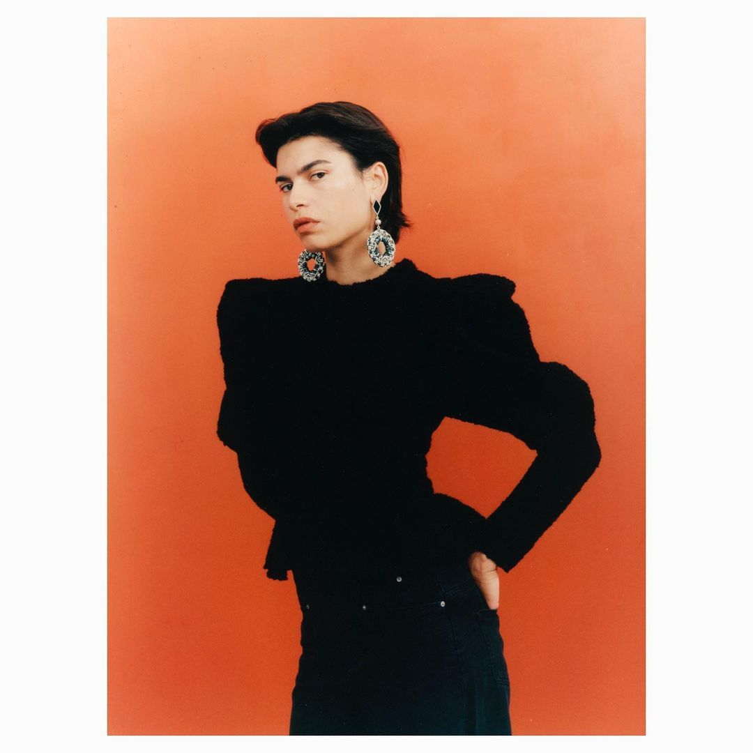 Portrait of a fashion model in a black top and dark pants, posing against an orange backdrop, showcasing modern earrings and style.