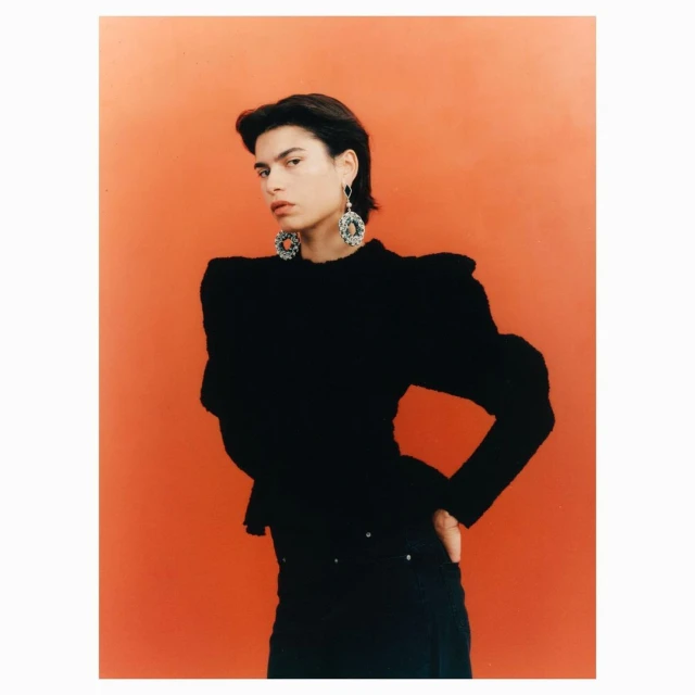 Portrait of a fashion model in a black top and dark pants, posing against an orange backdrop, showcasing modern earrings and style.
